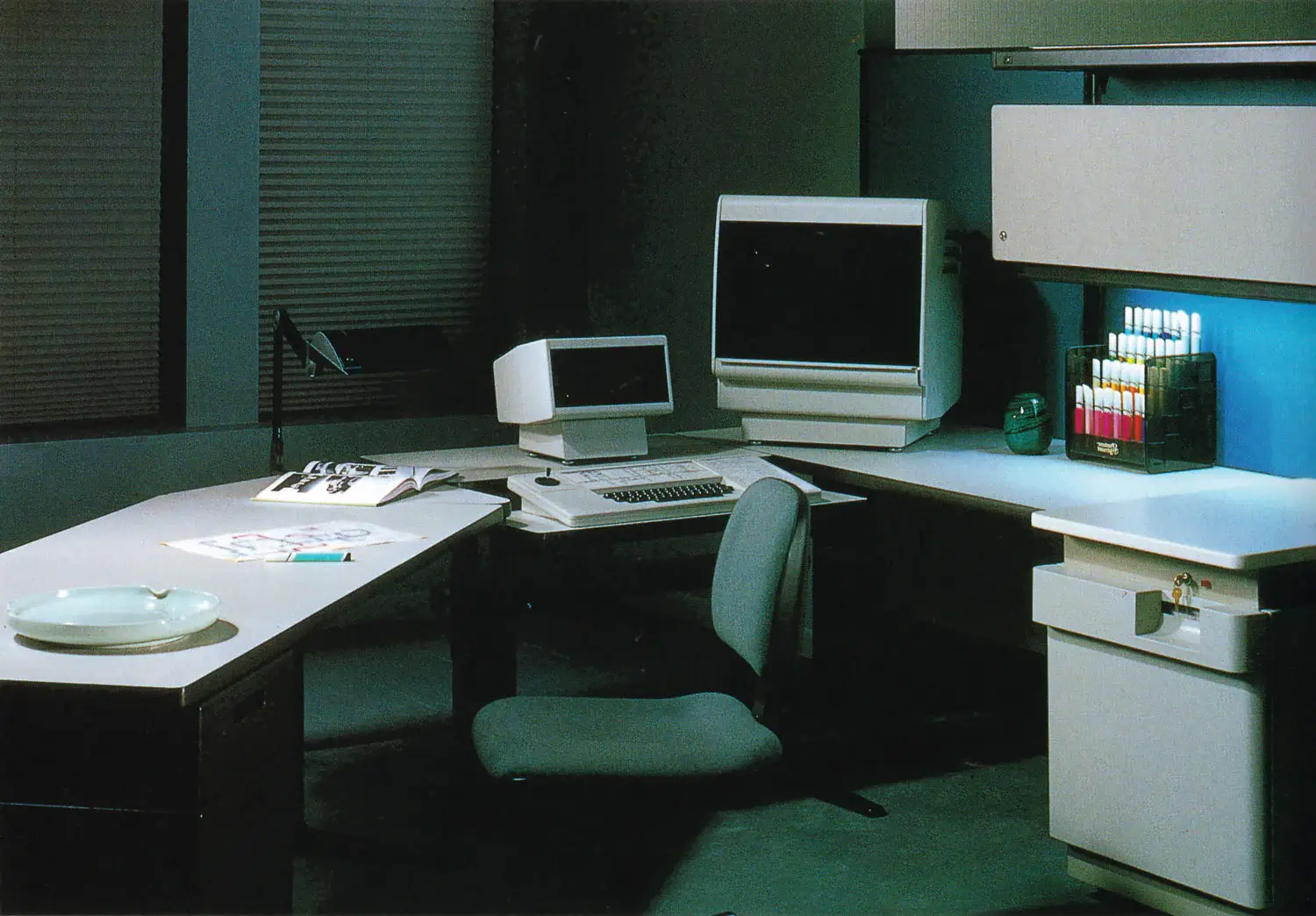 An office space from the late 1980s to early 1990s featuring an old-style desktop computer with a bulky monitor and separate keyboard. The computer is on a spacious desk that also holds an open book, some papers, and a drawing pad with a sketch and a marker on it. The room is dimly lit, primarily by a desk lamp, creating a quiet, studious atmosphere. It's pretty sick tbh.